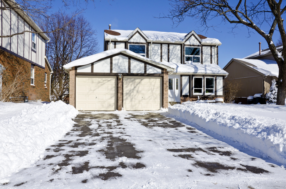 Two-story suburban house with dual garage and driveway, surrounded by snow and clear blue sky; snow is shoveled from driveway and walkway, and well-maintained trees hint at recent professional tree service.
