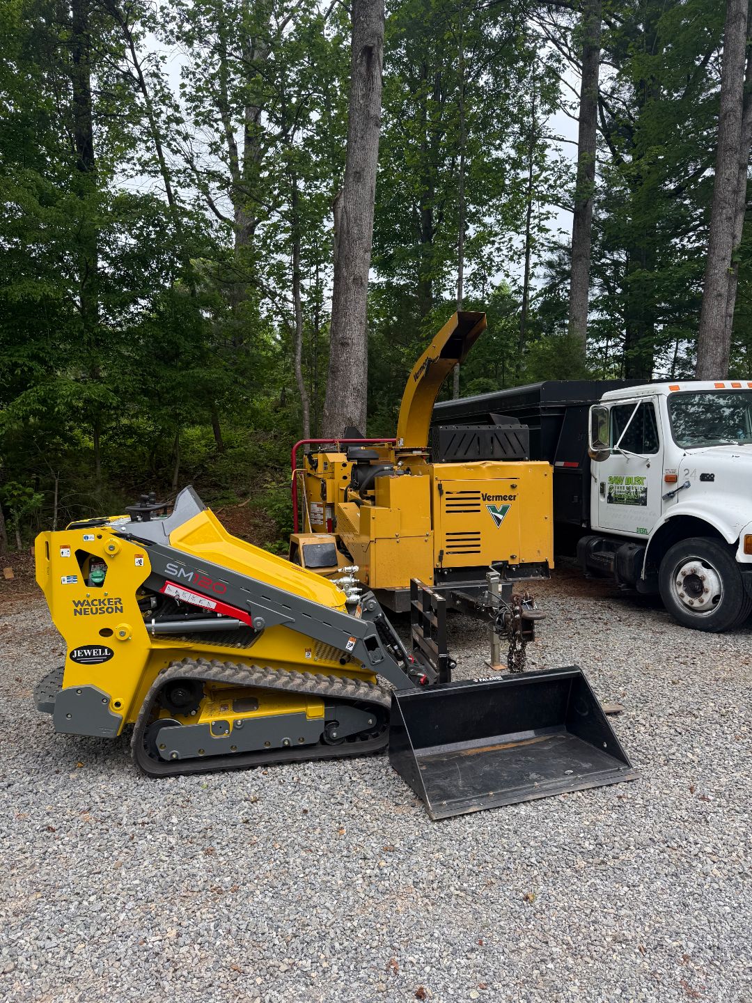 A yellow mini skid steer loader and a yellow wood chipper are parked on gravel beside a white truck in a wooded outdoor area.